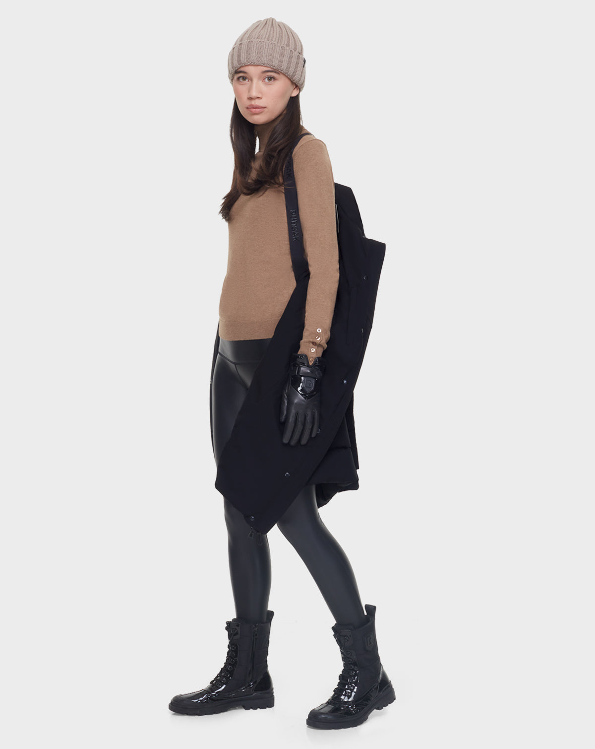 A woman stands against a plain white background holding the Rudsak ELISA - L121512 BLACK sustainable winter coat over her shoulder, dressed in a beige beanie, tan long-sleeve top, black gloves, shiny black leggings, and combat boots.