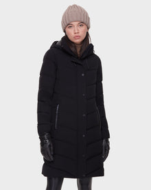 A woman wears the Rudsak ELISA - L121512 BLACK quilted down puffer, black leather gloves, black pants, and a beige knit beanie, standing against a plain light gray background with a neutral expression.