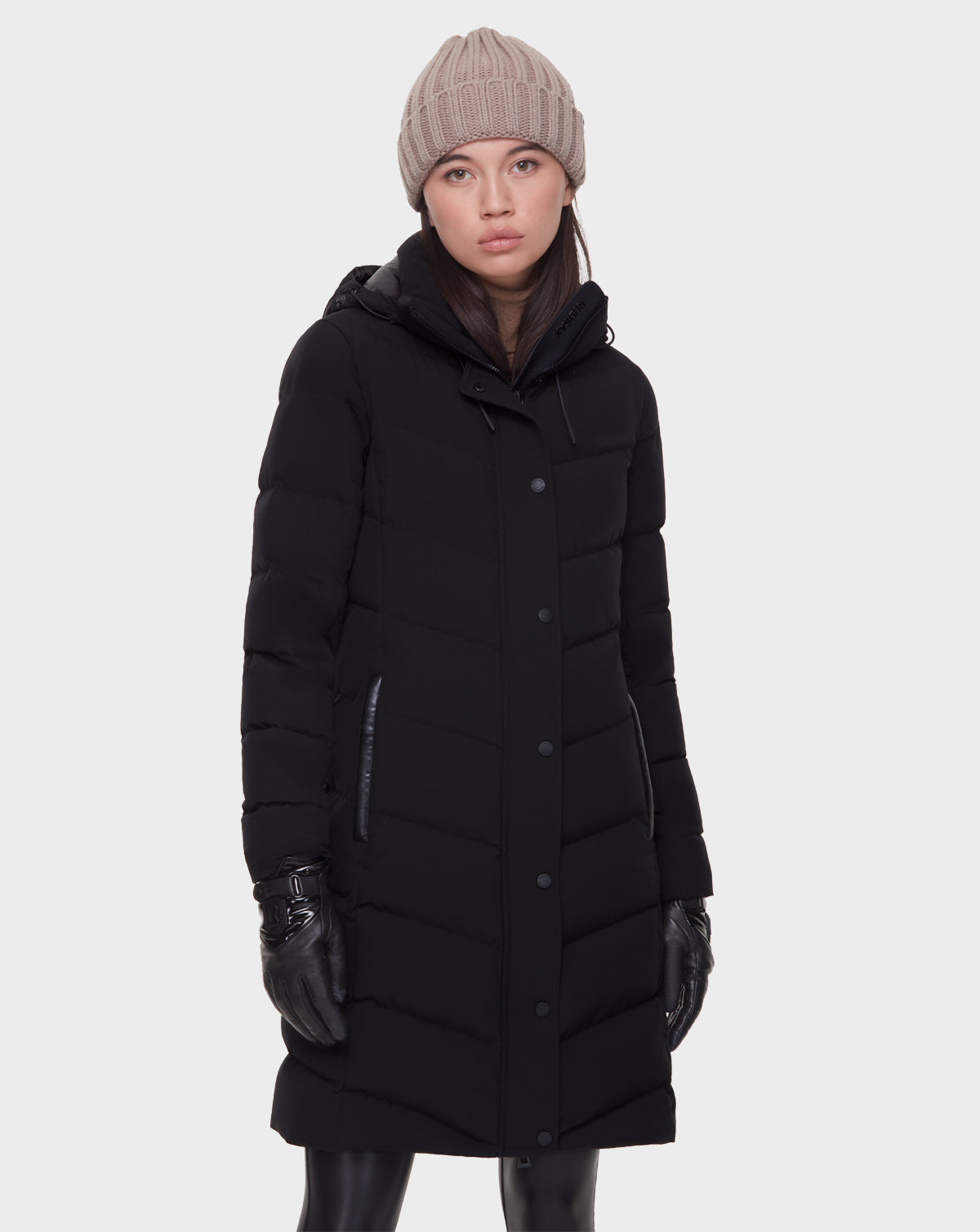 A woman wears the Rudsak ELISA - L121512 BLACK quilted down puffer, black leather gloves, black pants, and a beige knit beanie, standing against a plain light gray background with a neutral expression.