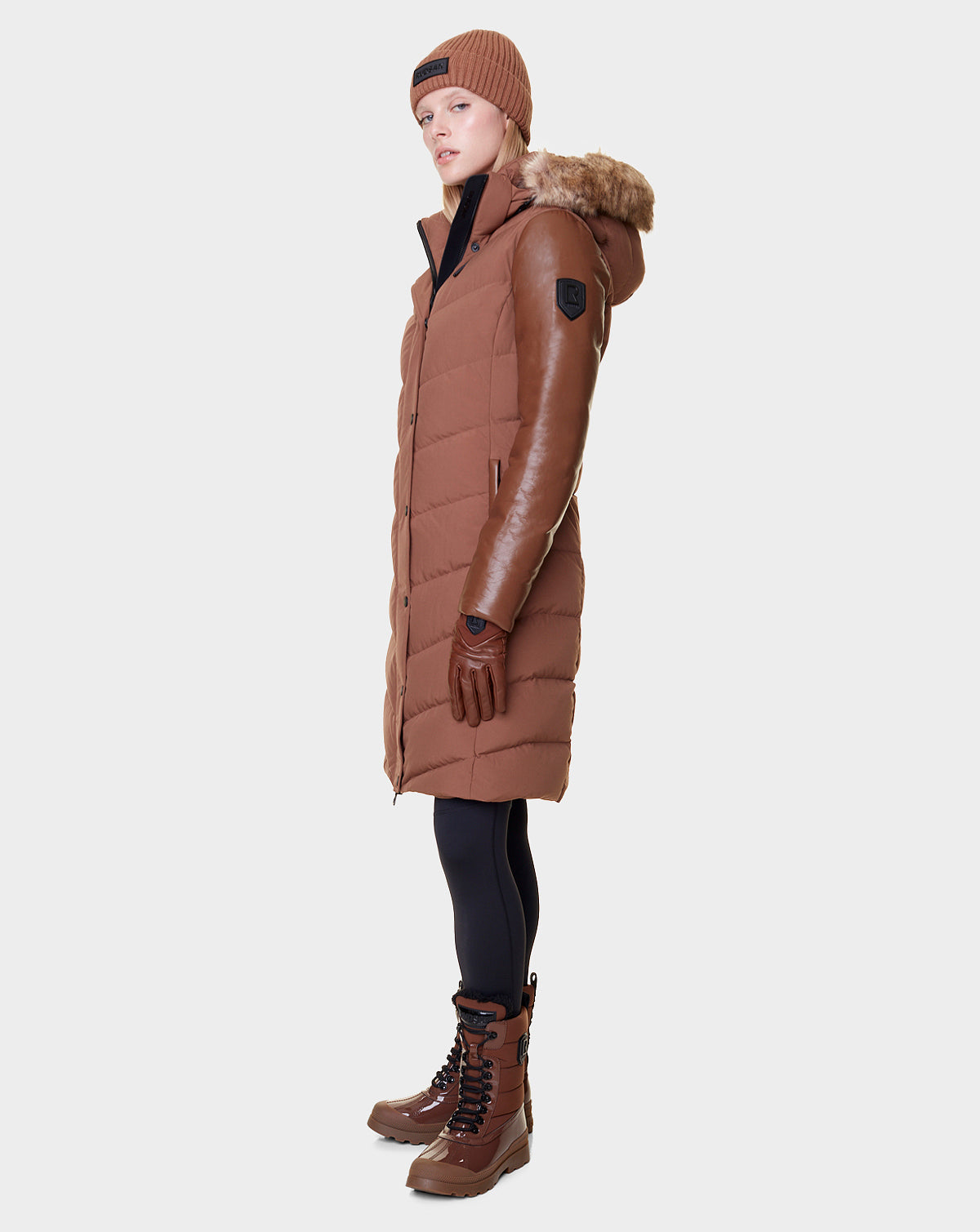 Wearing the Rudsak ELSA - L121519 TAUPE quilted down puffer, a person in brown gloves, knit hat, black leggings, and lace-up boots stands against a plain white background, turned slightly to the side.