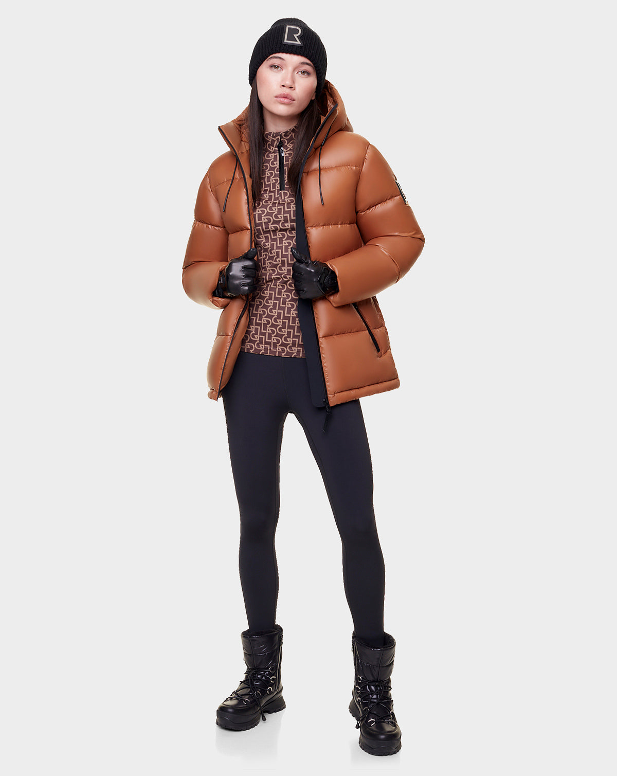 A woman stands facing forward against a plain white background, wearing the Rudsak EVIE - L122517 DARK CAMEL down puffer for women, a patterned brown top, black leggings, black winter boots, gloves, and a beanie.