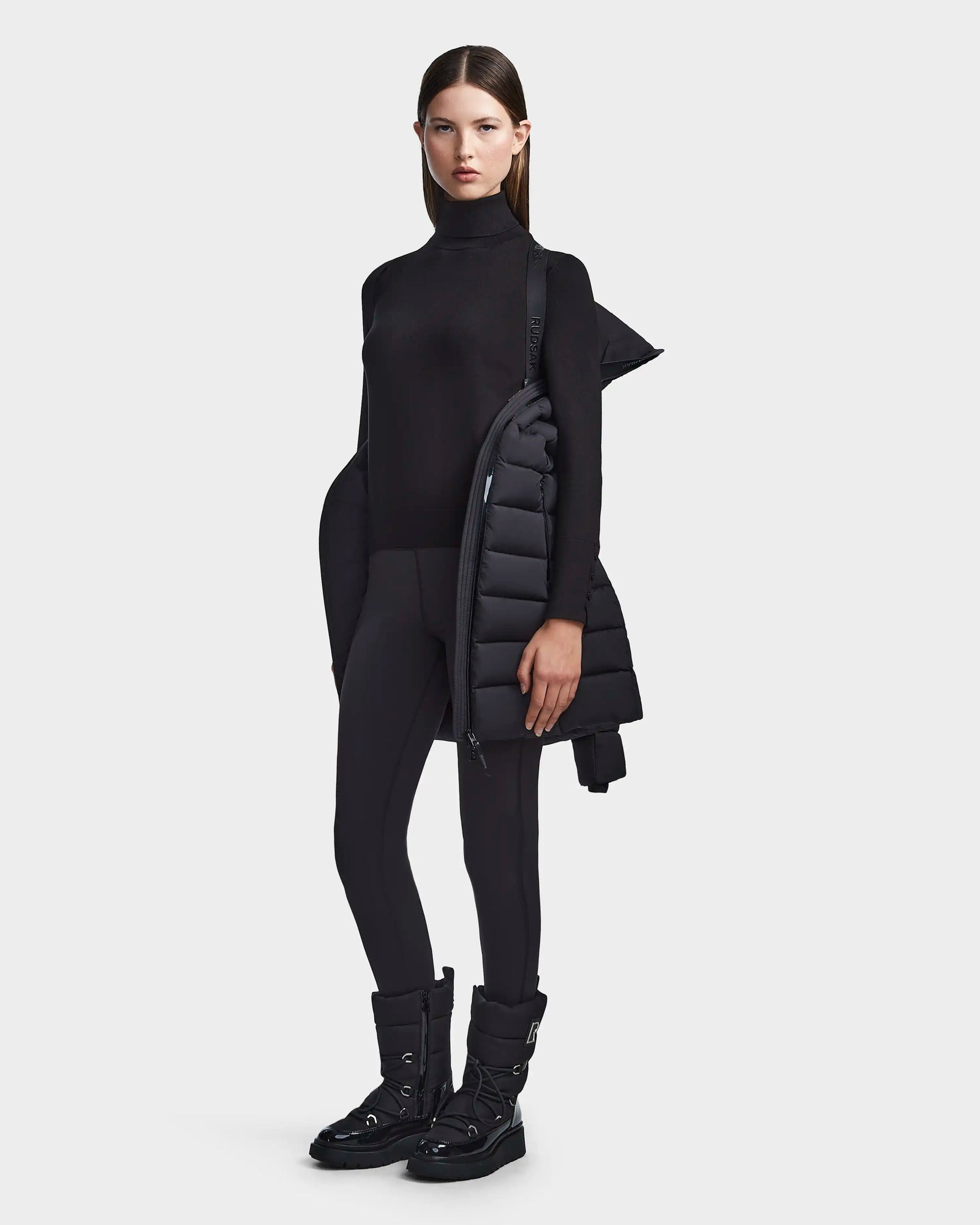 A woman stands against a light gray background wearing Rudsak's FRIDA M - L123514 Black women's mid-length down jacket with backpack-style straps draped over one shoulder, paired with a black turtleneck, pants, and winter boots.