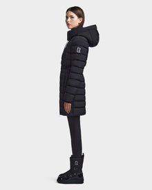A woman in profile wears the Rudsak FRIDA M - L123514 black mid-length down jacket with removable hood, black pants, and boots. The quilted jacket and boots display a white R logo. The background is plain and light.