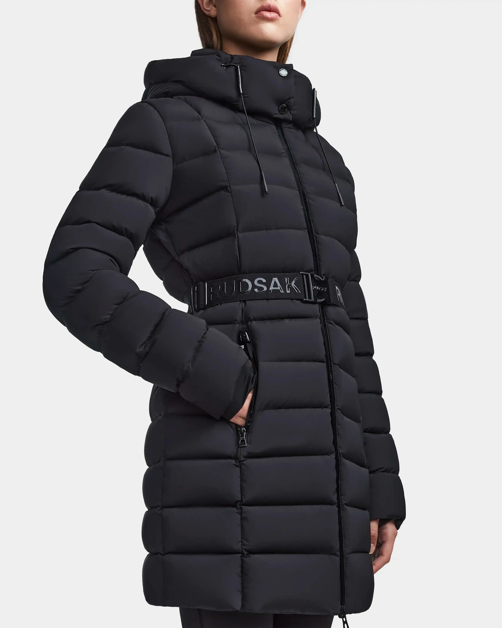 Wearing the Rudsak FRIDA M - L123514 Black, a women’s mid-length down puffer coat with a belted waist, front zipper, side pockets, horizontal stitching, and removable hood. The model’s face is partially out of frame.