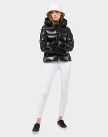 A person wears the Excluded MARE - 8121011 BLACK shiny black ultra-light jacket, a white bucket hat, white fitted pants, and black sneakers, standing with arms crossed against a plain light gray background.