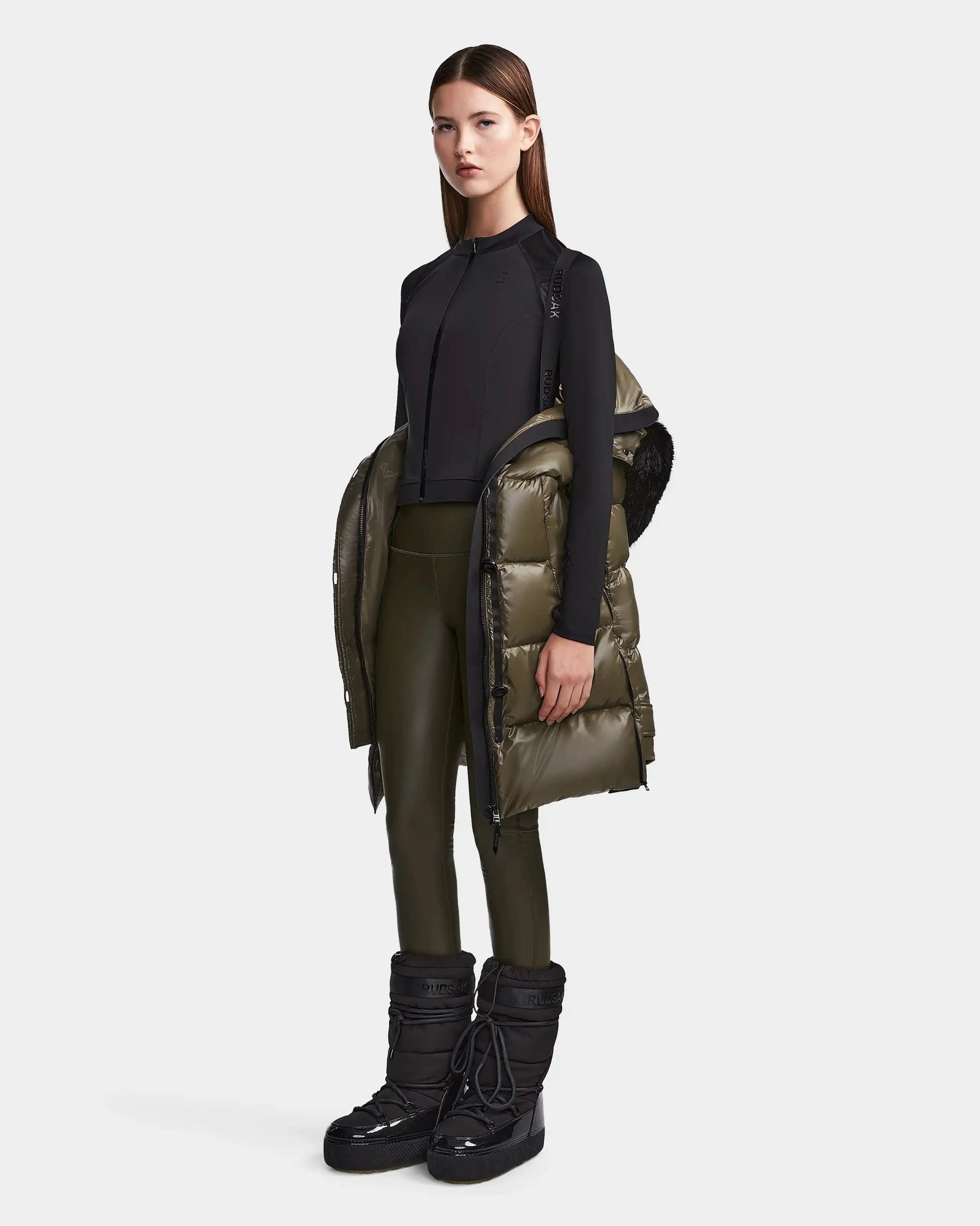 A woman stands against a light gray background, wearing the Rudsak MEYA S - L123521 Light Olive women's down puffer draped over her shoulders, paired with a black long-sleeve top, olive green leggings, and black winter boots.