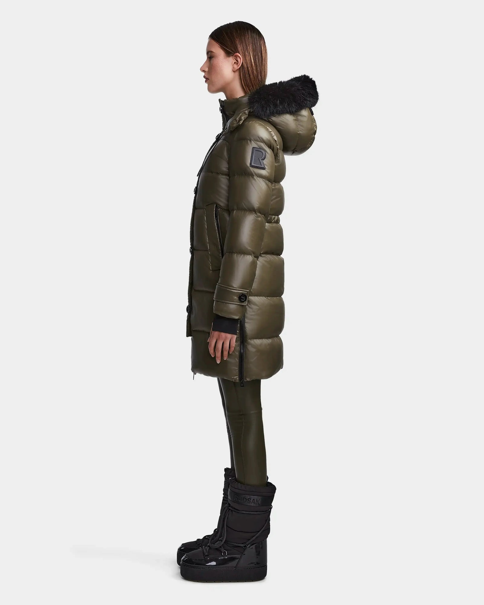 A woman stands in profile wearing the Rudsak MEYA S - L123521 Light Olive women's down puffer with a black fur-trimmed hood, paired with black pants and tall black boots against a plain white background.