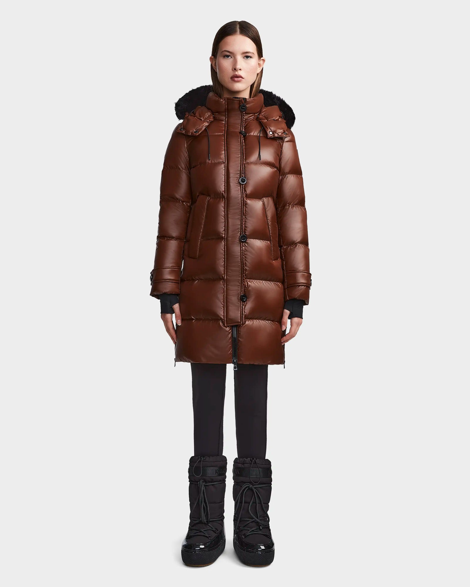 A woman stands facing forward in the Rudsak MEYA S - L123521 Cohiba down puffer with removable hood, styled with black leggings and winter boots, against a plain light gray background.