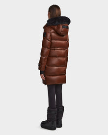 A person faces away, wearing the Rudsak MEYA S - L123521 Cohiba women's down puffer with a shiny brown finish, black fur-trimmed removable hood, paired with black leggings and winter boots against a plain light background.