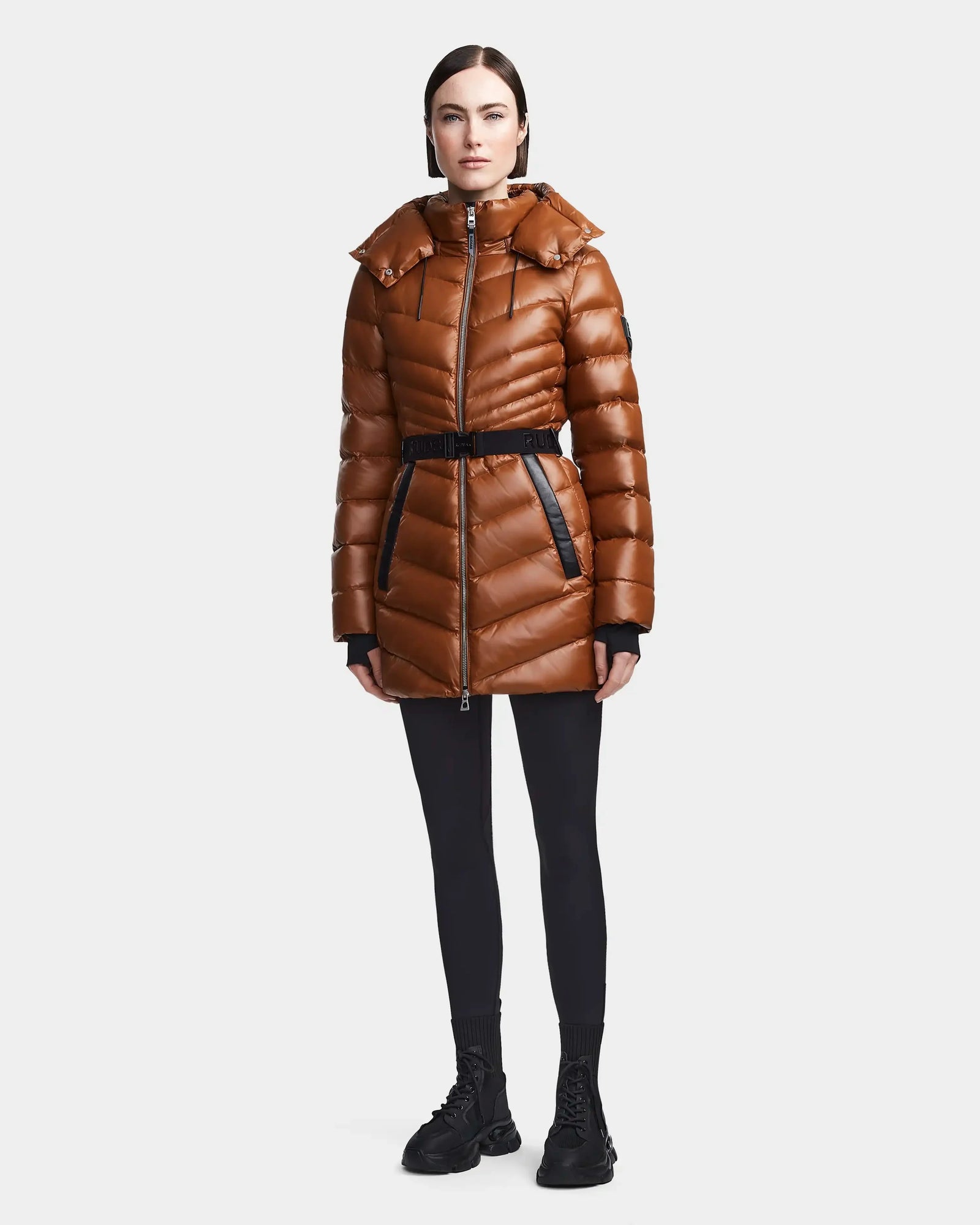 A woman faces forward wearing the Rudsak MALAYA - L123510 Dark Camel, a shiny brown women’s down puffer jacket with herringbone quilting, hood, and belt, paired with black leggings and sneakers against a plain white background.
