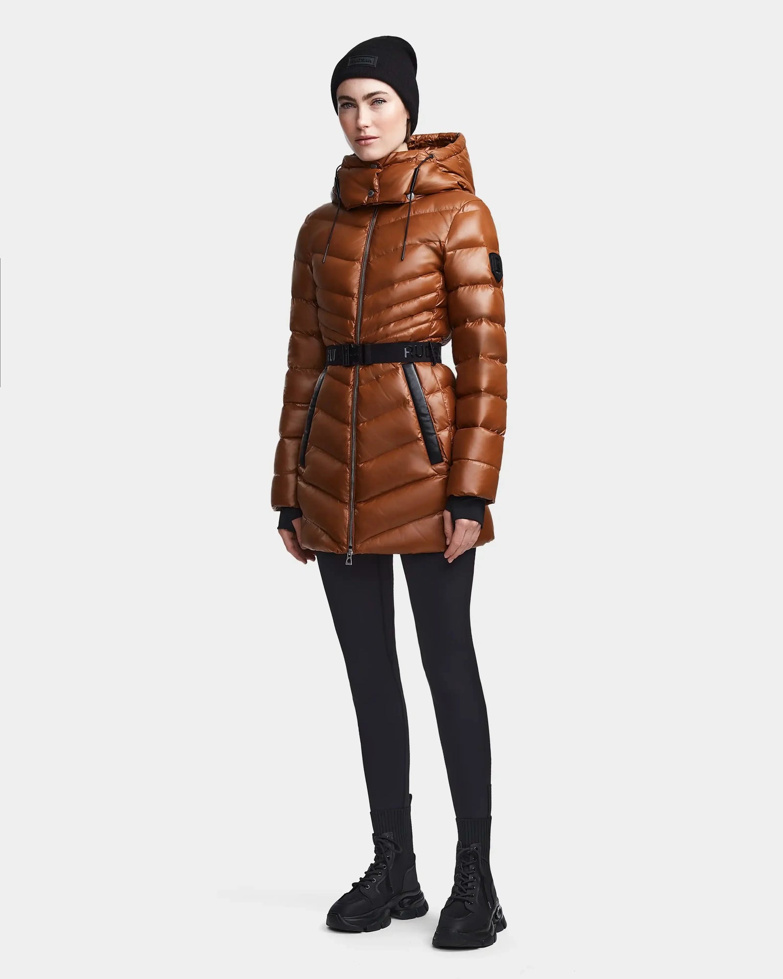 A person wears the Rudsak MALAYA - L123510 Dark Camel, a shiny brown quilted hooded coat with herringbone quilting, styled with a black belt, leggings, boots, and beanie against a plain white background.