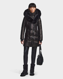 A person faces forward against a white background, wearing the Rudsak SHAUNA X - 8121568 BLACK women's winter coat with a large fur-trimmed hood and RDS down, paired with black leggings, boots, and holding a black quilted bag.