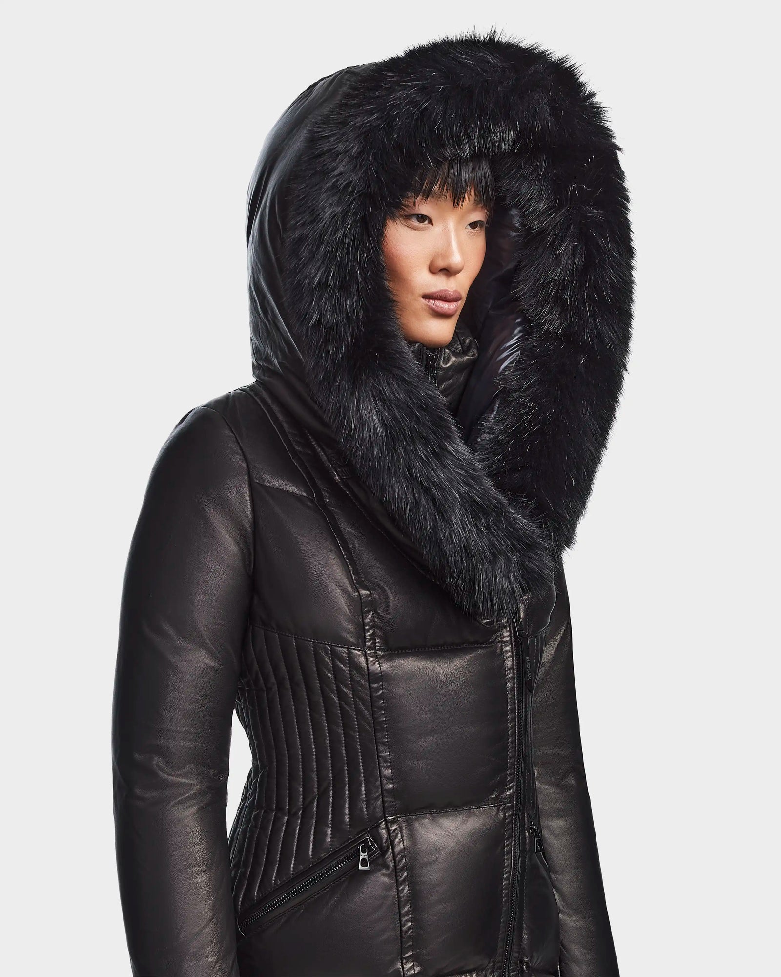 A woman in the Rudsak SHAUNA X - 8121568 BLACK puffer with a large, fur-lined hood stands slightly turned against a plain light background.