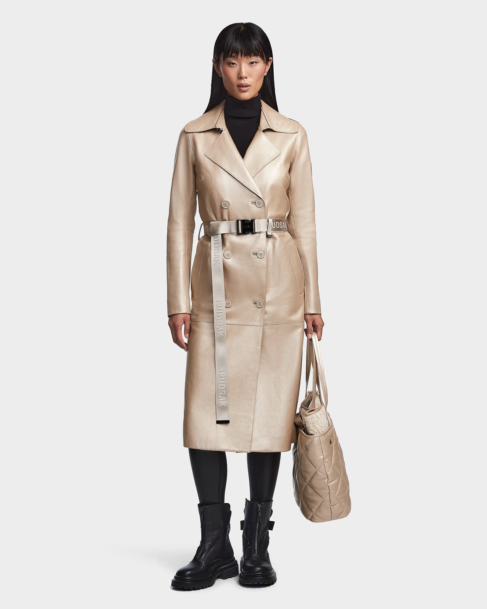 A woman stands facing forward, wearing the Rudsak KIRALY - 8123553 Metallic Ivory trench coat with a belted waist, black turtleneck, black leggings, black ankle boots, and holding a beige quilted tote bag.