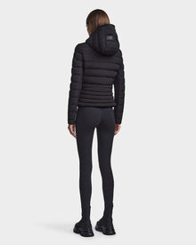 A person with straight brown hair, seen from behind, wears the RUDSAK MELISSA - 8122551 BLACK womens puffer jacket, black leggings, and black boots against a plain white background.