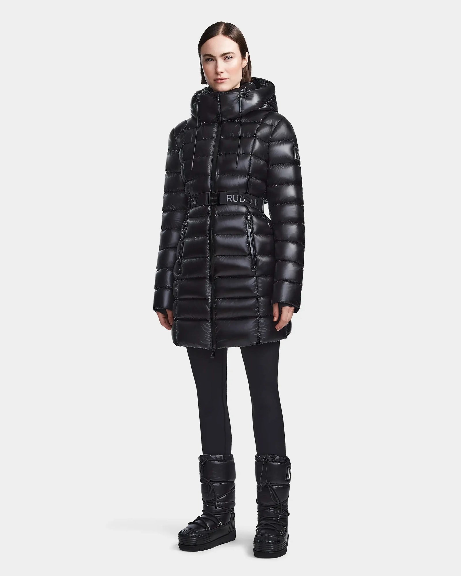 A woman in the Rudsak FRIDA S - L123513 Black mid-length down jacket with removable hood, black leggings, and matching puffer boots stands against a plain white background.