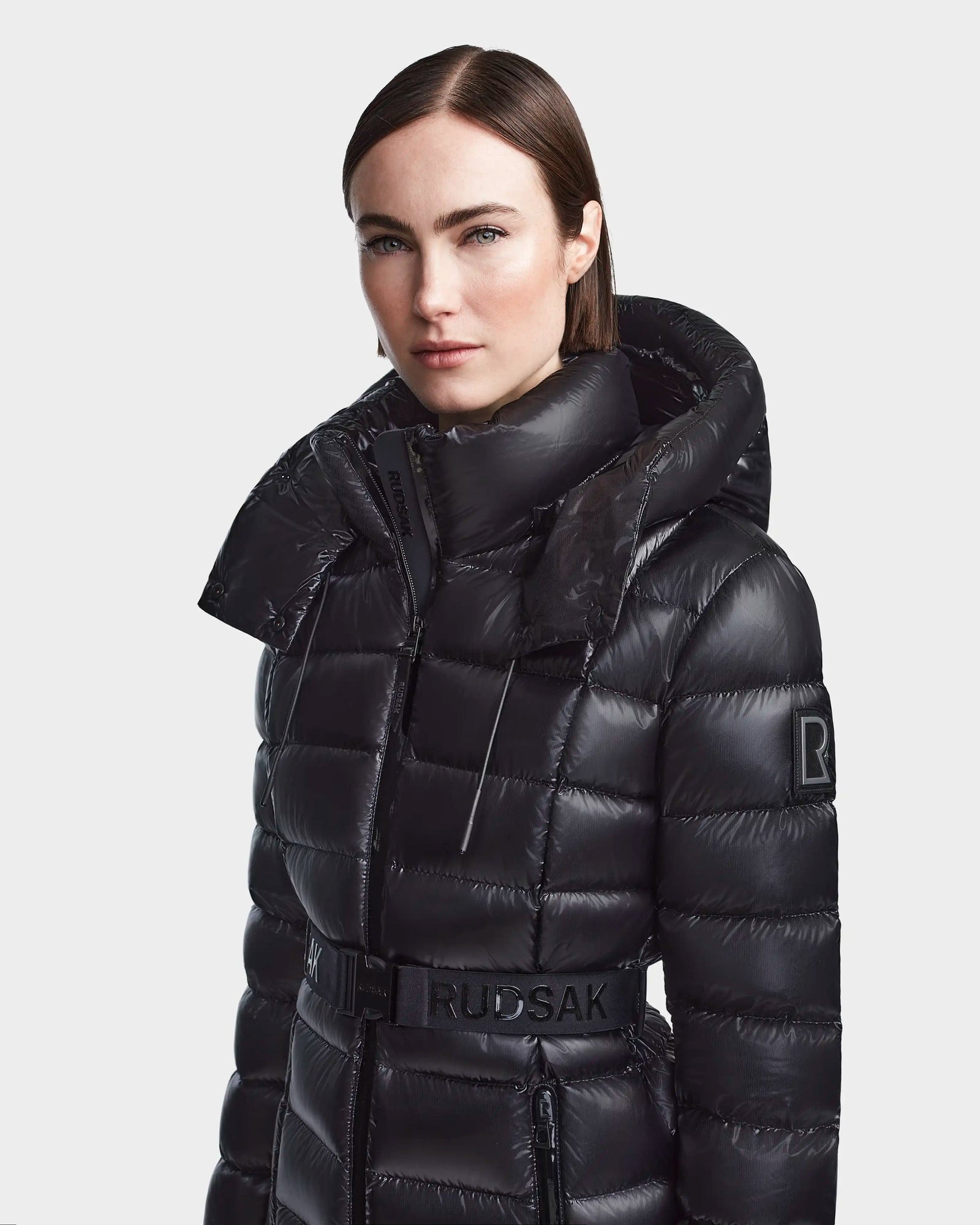 A woman with straight brown hair wears the Rudsak FRIDA S - L123513 Black mid-length shiny quilted down jacket with a high collar, removable hood, belted waist featuring RUDSAK branding, and an R patch on the sleeve, set against a white background.