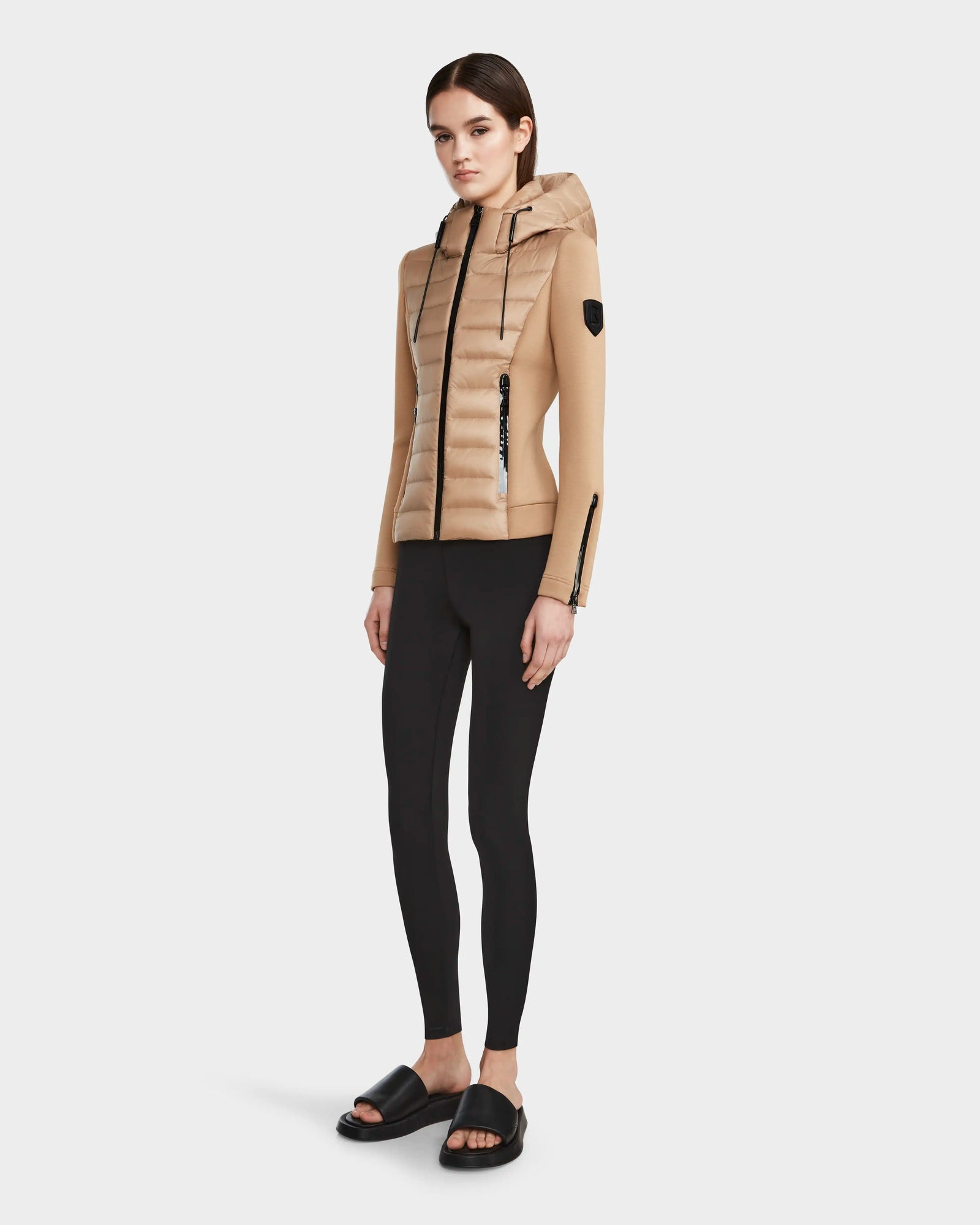 A woman faces forward, wearing the RUDSAK MABS - 8122020 TOFFEE lightweight jacket with waterproof zippers, black leggings, and black slide sandals against a plain light gray background.