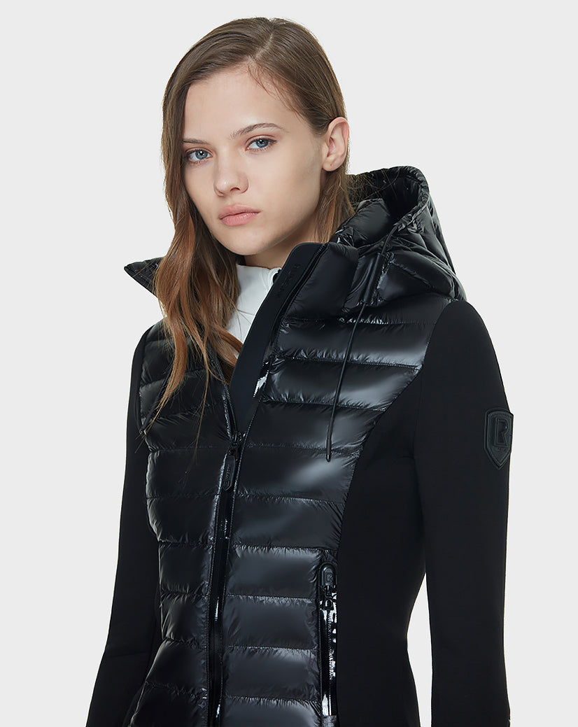 A young woman with long, light brown hair wears the Rudsak MABS - 8121012 BLACK puffer vest, featuring waterproof zippers and a hood, over a black long-sleeve top. She stands against a plain light gray background, looking forward neutrally.