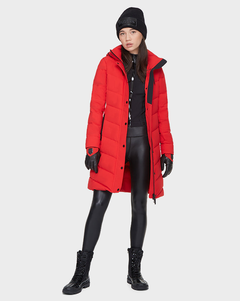 A woman in the Rudsak ELISA - L121512 POPPY RED quilted down puffer, black leggings, lace-up boots, gloves, and a beanie stands against a plain light background.