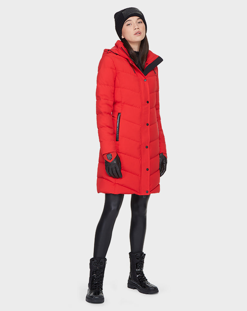 A person stands against a white background wearing the Rudsak ELISA - L121512 POPPY RED women’s quilted down puffer coat, filled with RDS-certified down for sustainable warmth, paired with a black beanie, gloves, leggings, and lace-up boots.