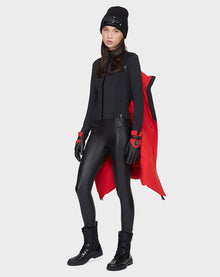 A woman poses against a light backdrop, draping the Rudsak ELISA - L121512 POPPY RED sustainable winter jacket over her shoulder, styled with a black beanie, long-sleeve top, faux leather leggings, gloves, and boots for a sleek look.