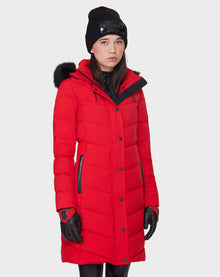 A woman wearing the Rudsak ELISA - L121512 POPPY RED quilted down puffer with a fur-lined hood, paired with black gloves and a black beanie, stands against a plain background—a stylish choice for winter warmth.