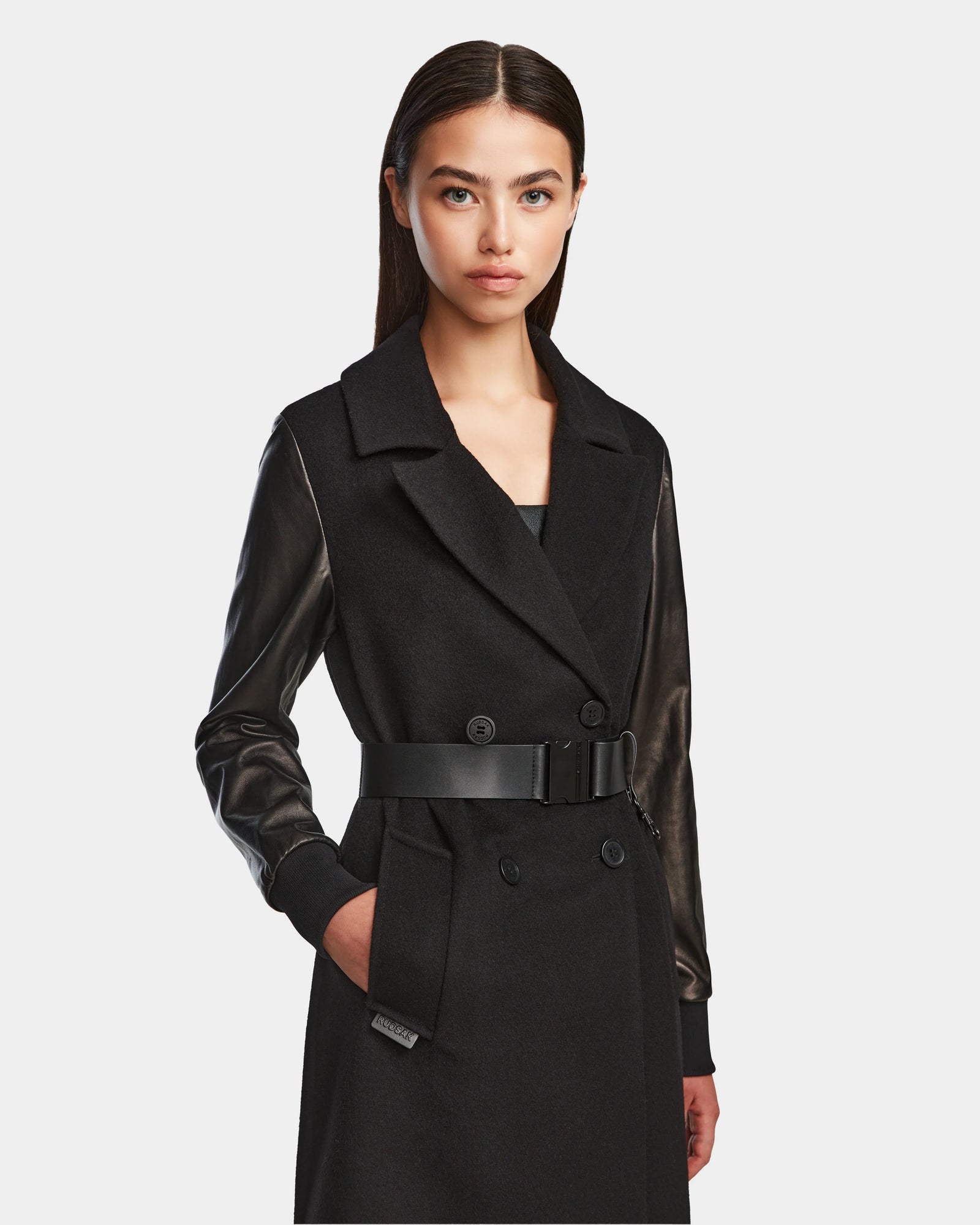 A woman with straight dark hair models the Rudsak MARYLIN - 8124520 Black women’s wool coat with leather sleeves, a wide collar, and a belted waist. She stands facing forward, her right hand in her pocket against a plain light background.