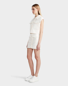 A woman with straight blonde hair stands sideways in the Rudsak SOLSA - 8124048 Eggshell womens cropped quilted vest with eco-friendly Sorona, layered over a sleeveless white top and matching skirt, paired with white sneakers against a plain background.