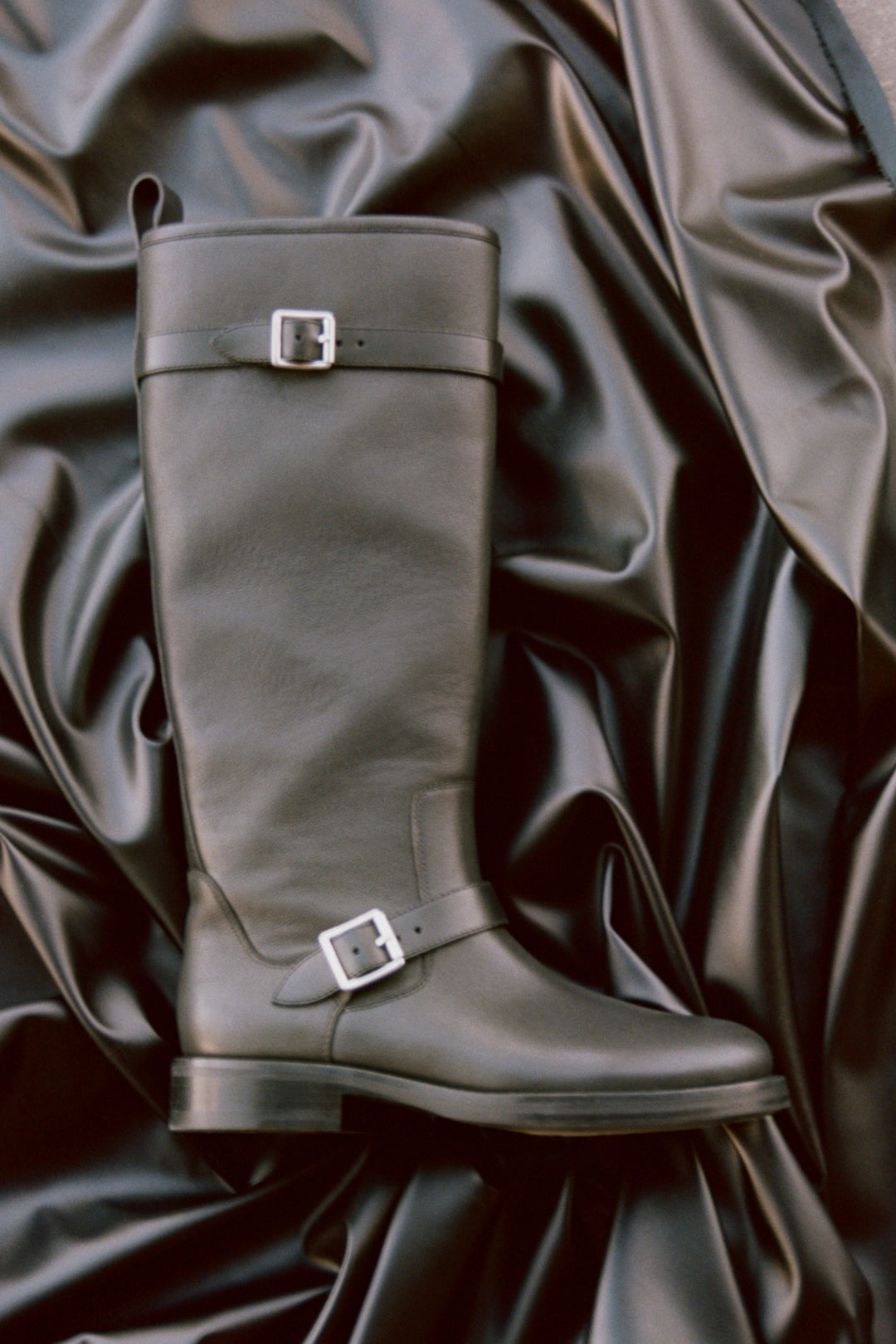 Gray leather knee-high boot with metal buckle straps, displayed on draped fabric background.