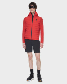 A person wearing a RUDSAK JARRY - 6122007 POPPY RED down hooded puffer jacket, black shorts, black socks, sneakers, and large black sunglasses stands against a plain white background.