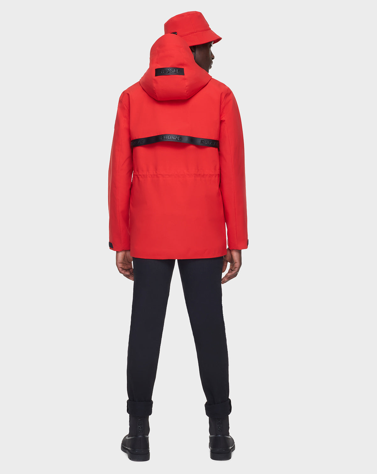 A person wears the RUDSAK JACY - 6122000 POPPY RED hooded jacket and a matching red bucket hat, paired with black pants, boots, and a black branded strap, standing against a light gray background.