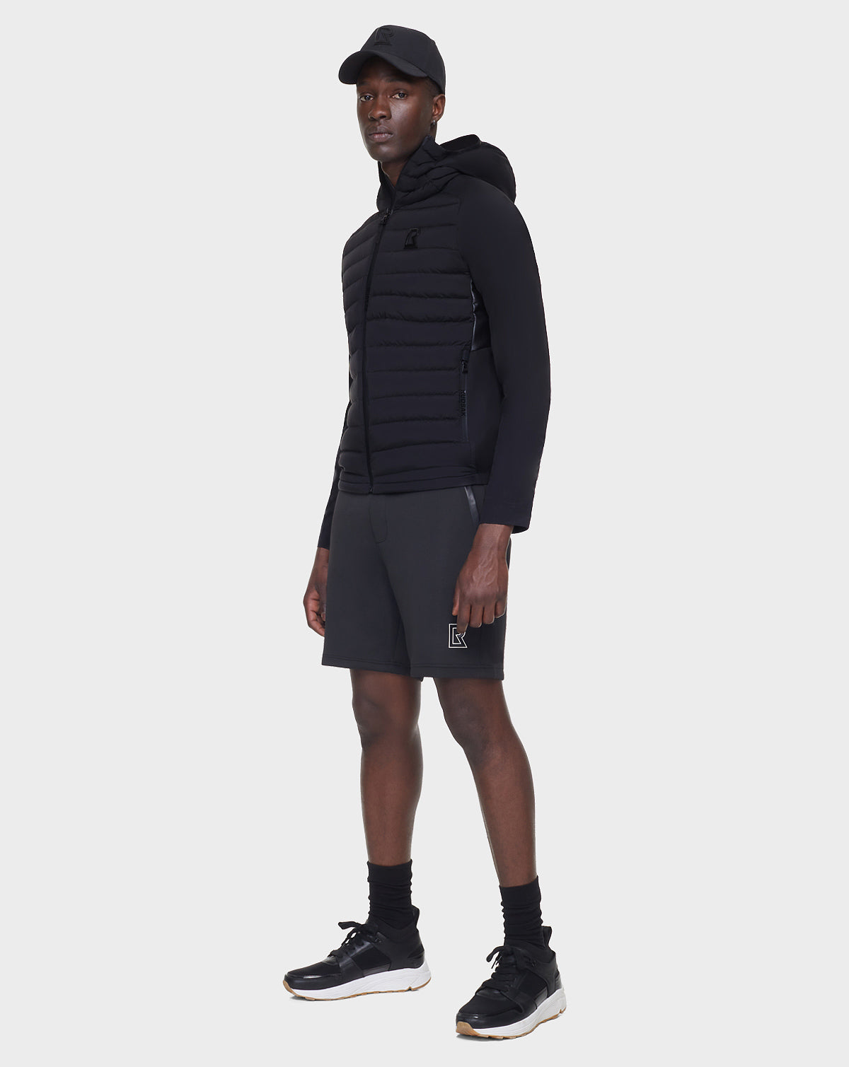 A man wears the RUDSAK JARRY - 6122007 BLACK RDS-certified down hooded jacket, paired with a black cap, shorts, socks, and sneakers, standing against a plain light background.