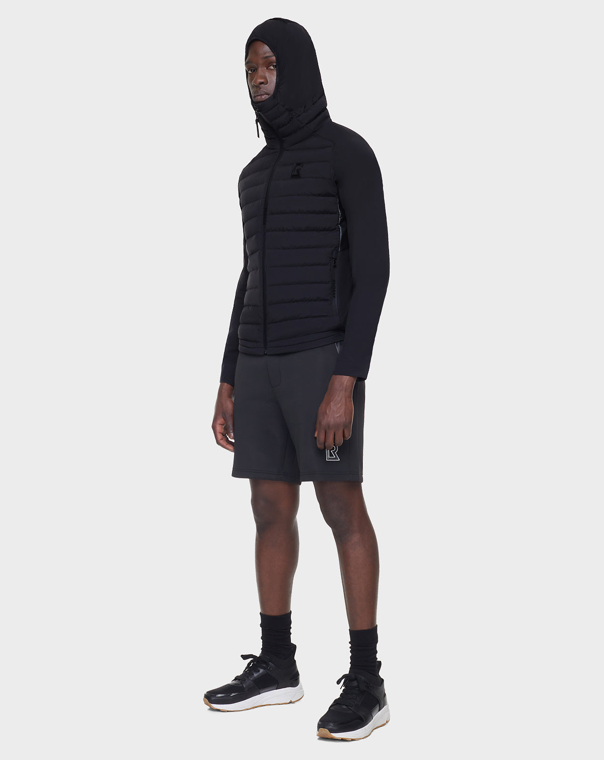 A person wearing the RUDSAK JARRY - 6122007 BLACK men’s windbreaker, along with black shorts, socks, and sneakers, stands against a plain white background facing slightly to the side.