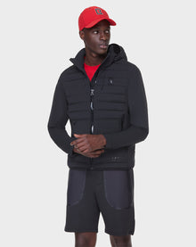 A young man in the RUDSAK JAS - 6122006 BLACK hooded puffer jacket with RDS-certified down, a red cap and shirt, and black shorts stands with hands in his pockets, looking to the side against a plain white background.