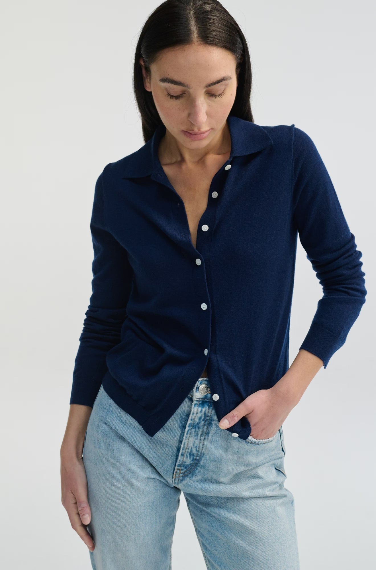 Woman wearing a navy blue cardigan over a white shirt with light blue jeans on a plain background