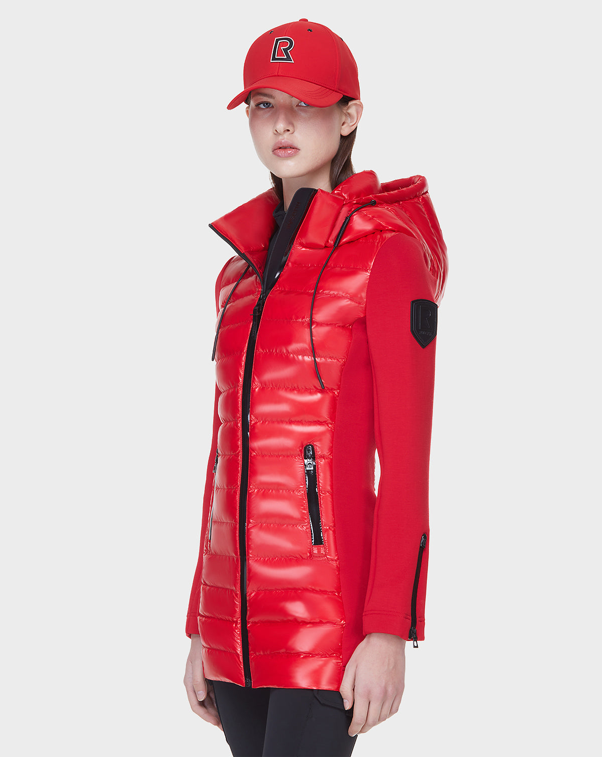 A person wearing the Rudsak MERINA - 8121006 POPPY RED women’s light jacket stands against a plain white background, styled with a red cap featuring a black R logo and black pants.