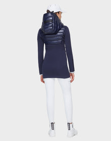 A woman faces away, wearing the Rudsak MARRY - CA EN L121003 DEEP BLUE puffer jacket with shiny hood, paired with white leggings, sneakers, and cap. The activewear-inspired look stands out against a plain light gray background.