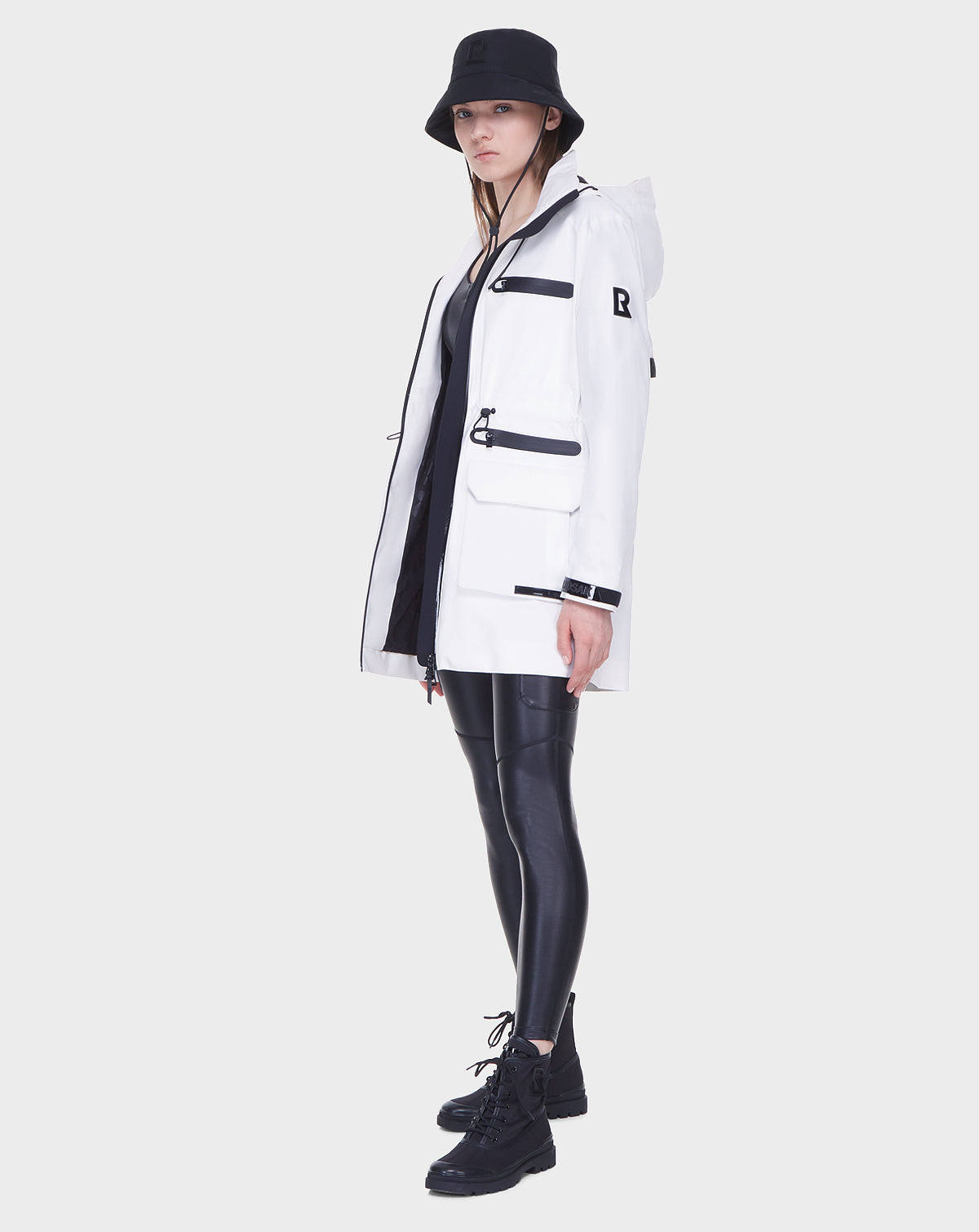 A person wearing the Rudsak CAMY - 8122002 WHITE mid-length raincoat stands against a plain white background, styled with a black bucket hat, shiny black leggings, and black lace-up boots.