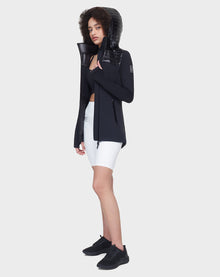 A woman with curly hair wears the Rudsak CAMIRA - 8122011 BLACK activewear jacket with waterproof zippers over a black sports bra, white biker shorts, and black sneakers, standing against a white background and looking ahead neutrally.