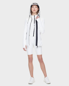 A woman wears the Rudsak CAMIRA - 8122011 WHITE activewear jacket with a hood, paired with white cycling shorts and sneakers. She holds the hood, ready for outdoor workouts against a plain light gray background.